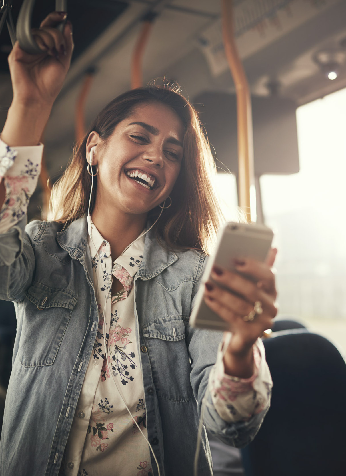 Young woman wearing earphones laughing at a text message on her cellphone while riding on a bus